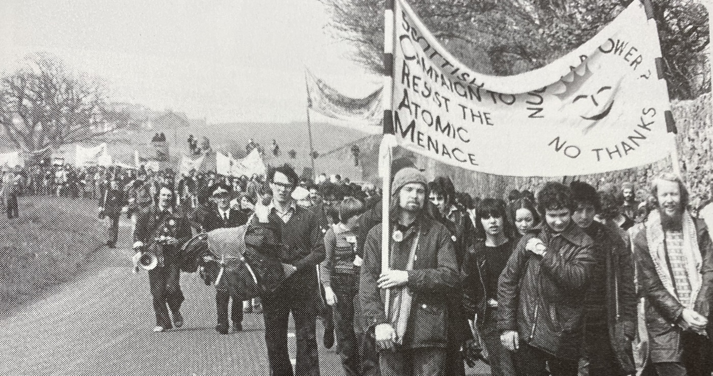 A black and white photo of a demonstration with people carrying a banner that says 'Scottish Campaign to Resist the Atomic Menace' and 'Nuclear Power? No Thanks' with a sun with a smiley face in middle. Source: 'Torness: Nuclear Power Station: From Folly to Fiasco' (Edinburgh: SCRAM, 1983)
