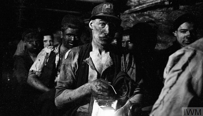A black and white photo group of miners underground, walking forward led by a serious older man with a moustache wearing a helmet and holding a lamp. The face of the man behind is completely black with coal dust.Source: Ministry of Information, 1942, Imperial War Museum https://www.iwm.org.uk/collections/item/object/205199325