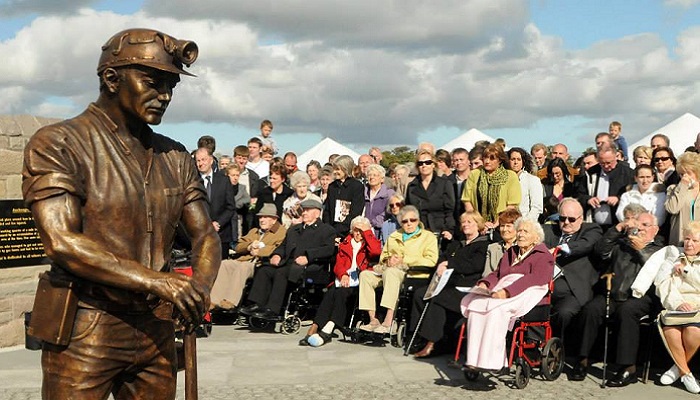 A statue of a miner leaning on a pickaxe with a crowd of people including older people in wheelchairs around it. Source / Copyright: https://www.facebook.com/Auchengeich-Miners-Memorial-916368968418660/photos
