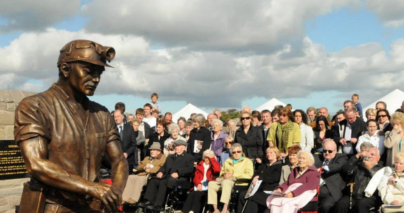 A statue of a miner leaning on a pickaxe with a crowd of people including older people in wheelchairs around it. Source / Copyright: https://www.facebook.com/Auchengeich-Miners-Memorial-916368968418660/photos
