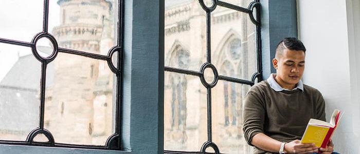 A young man sitting on an window sill in an old University building reading a book. Source: Glasgow University Image Library