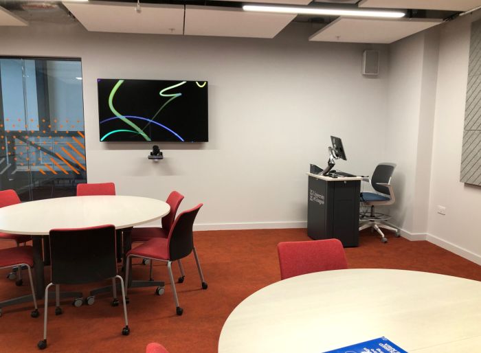 Flat floored teaching room with tables and chairs, PC, lectern, and large screen.