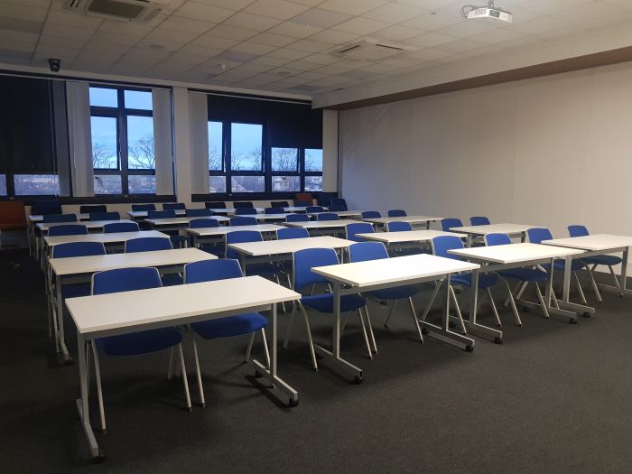 Flat floored teaching room with rows of tables and chairs, and projector.