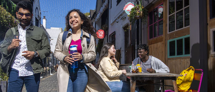 Two students walking along in an old-fashioned lane in Glasgow with two more students sitting a table in the sunshine behind them. Source: GU Image Library 