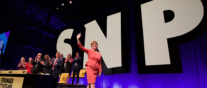 Nicola Sturgeon waving at a crowd at an SNP conference with panellists clapping behind her and a SNP logo behind her on stage Source: Terry Murden Publisher:Shutterstock Link: https://enterprise.shutterstock.com/catalog/licenses