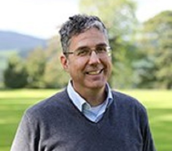 An image of Fabric Ramound outside in a forest wearing a grey jumper and a blue shirt