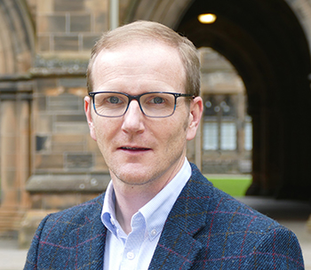 Image of Professor Graeme Roy wearing a blue shirt and dark blue blazer standing with the University Cloister behind him