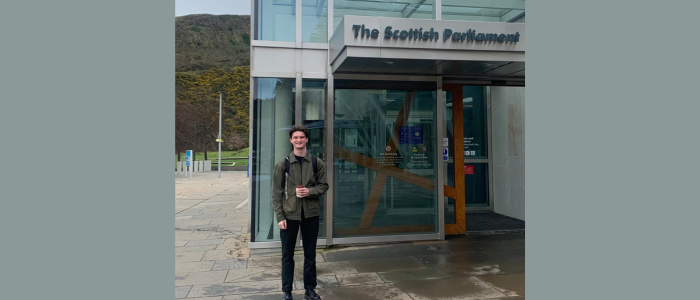 Young male wearing a dark jacket and trousers standing in front of the Scottish Parliament Building 