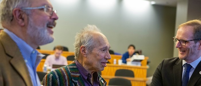 Sam Peltzman with Graeme Roy and another person beside them chatting in the centre of a lecture hall. Close-up on Sam Peltzman's animated face and raised hands while he is speaking to a person whose face can't be seen. Source: Chicago Booth University