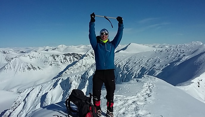 A student stood atop a snow-capped mountain in Sweden holding his pick axe above his head with two hands