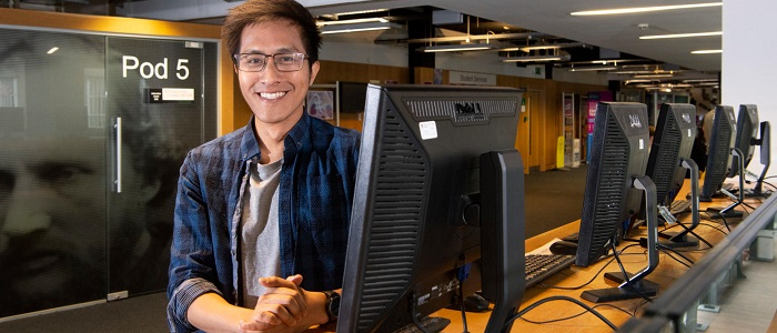 A staff member stood beside a PC in the library, with hands clasped and smiling at the camera
