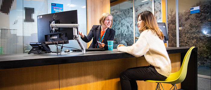A staff member sat on one side of a desk gesturing towards a monitor whilst advising a student