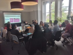 A group sitting round a table with a slide on display on a large screen in the background
